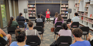 A small crowd of students listen to a woman reciting poetry.