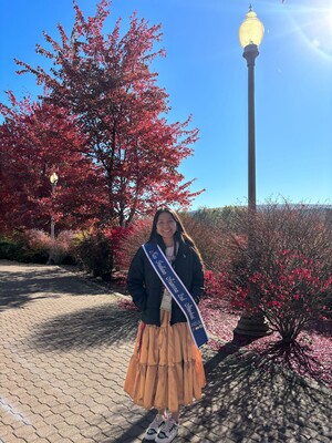Photo of Tonana Ben, Library Aide at Labriola Center. She is standing in front of red bushes and a red tree wearing a long yellow skirt with a sash across her shoulder that says "Miss Indian Arizona 2nd attendent" 