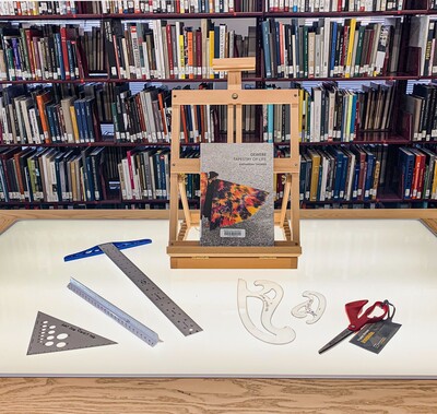 A display of tools available for lending. Left to right is a triangle, ruler, t-square, easel, S curves, and left-handed scissors.